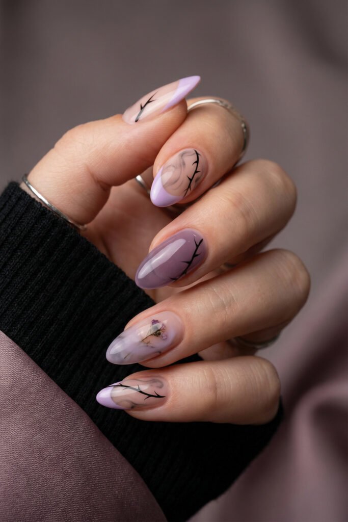 Moody lavender edgy spring nails with smoky airbrushed details, black branch art, and a delicate dried flower accent on almond nails.
