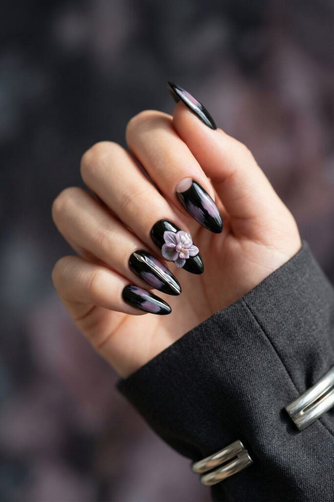 Black almond nails with iridescent purple accents and a 3D lilac flower, blending glossy drama with floral softness in a spring goth nails look.
