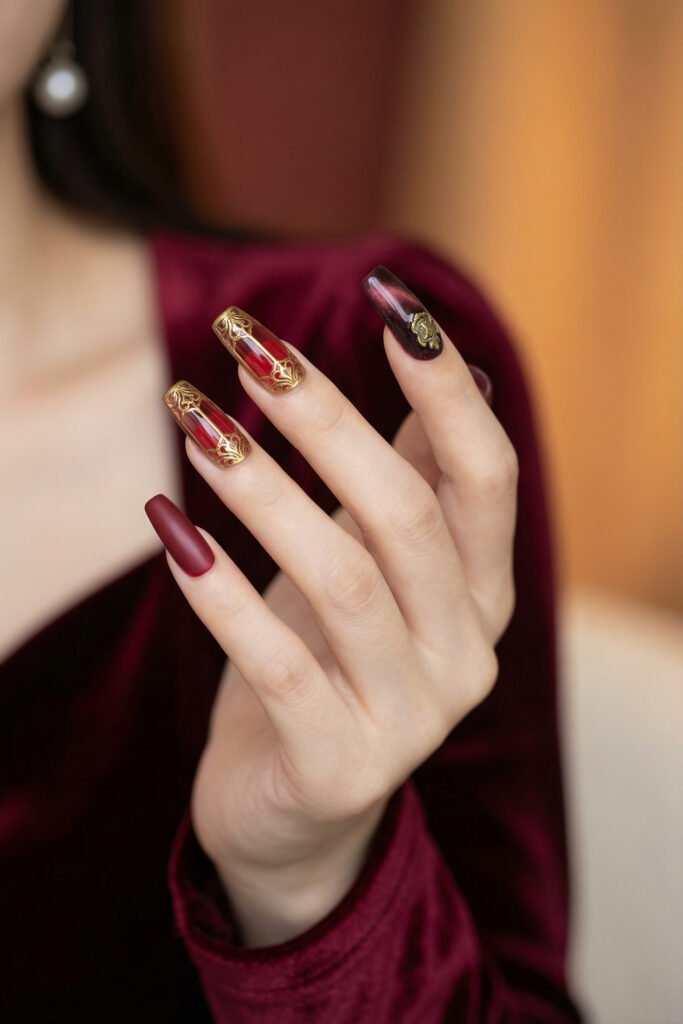 Chic Gold and Red Nails for a Bold, Polished Look 7 Hand with burgundy coffin nails featuring ornate gold filigree accents and deep wine details with a velvet sleeve against a softly lit background