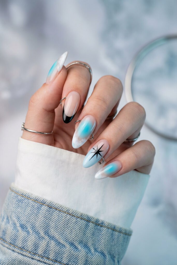 Icy blue and white edgy spring nails with black French tip accents, soft aura airbrushing, and a sharp black star design on almond nails.
