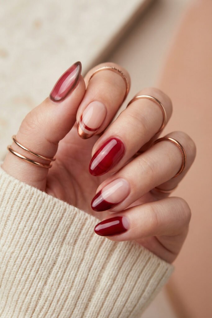 Glossy almond fall red nails with sheer cranberry ombré, curved copper French accents, deep red polish, and nude negative-space details.
