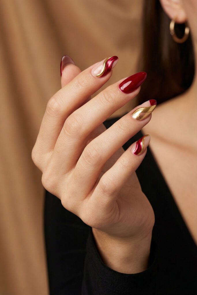 Chic Gold and Red Nails for a Bold, Polished Look 16 Close up of a hand with red and nude almond nails featuring flowing gold swirls and one mirror gold accent nail against a warm blurred background