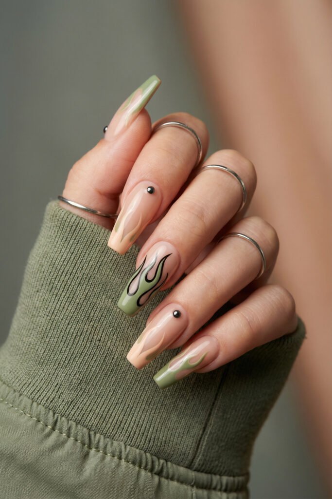 Glossy edgy spring nails with green flame tips, black outlined details, nude base, and small stud accents on square nails.

