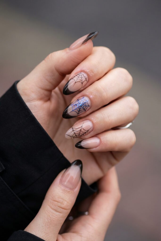 Nude almond nails with black ombré French tips and spider web art, including one shimmering blue web accent for chic spring goth nails.
